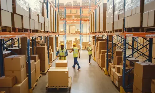 Magazijnmedewerkers in veiligheidshesjes lopen tussen hoge stellingen met dozen